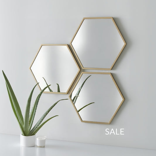 Three hexagonal mirrors with gold frames on a white wall, with a plant and books in the foreground.