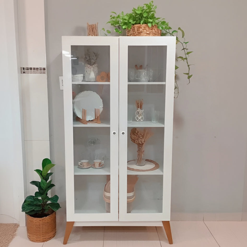 White glass-fronted cabinet with wooden legs displaying decorative items against a light wall.